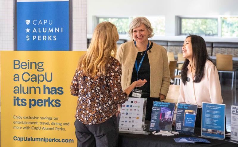 An attendee speaking with CapU Alumni staff members at the CapU Alumni Perks display table during an on-campus event.