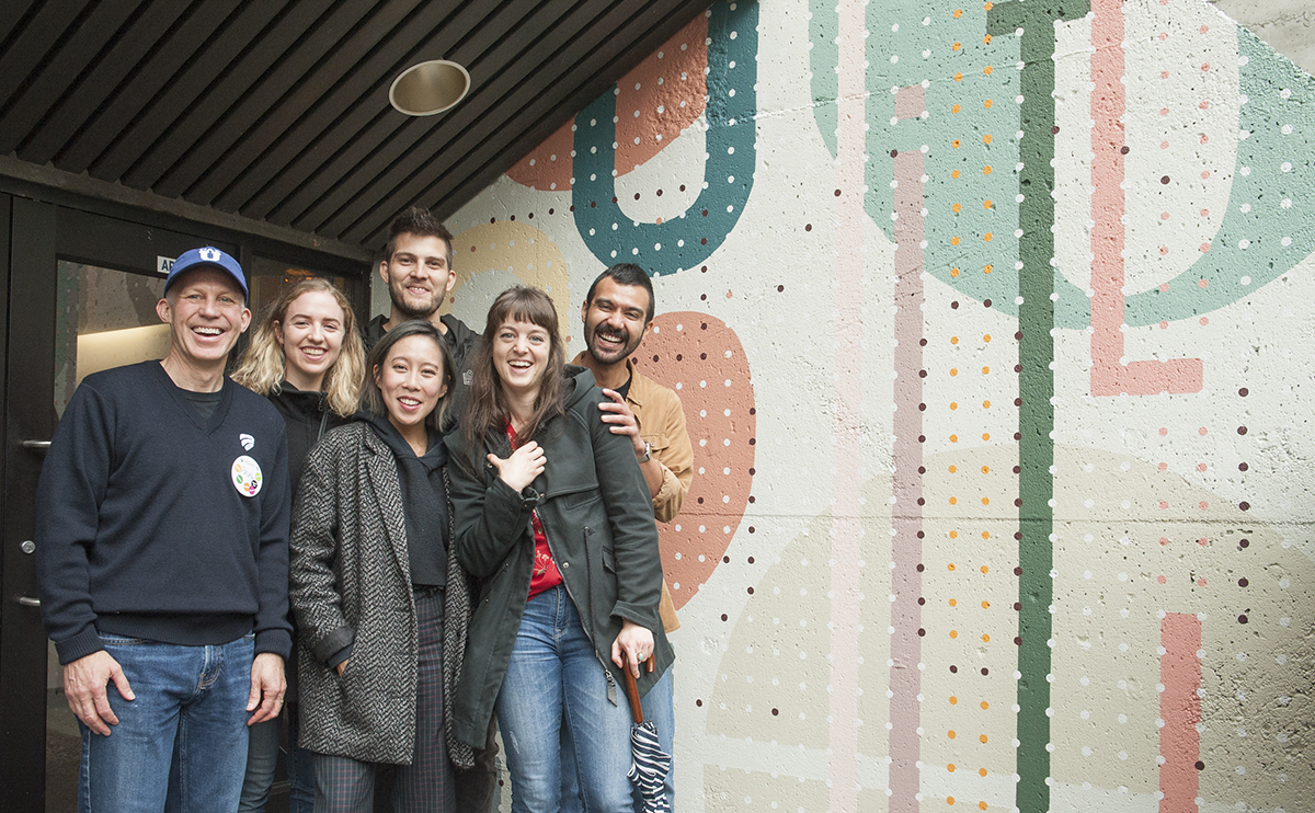David Vertesi smiling with students and CapU President Paul Dangerfield in front of mural