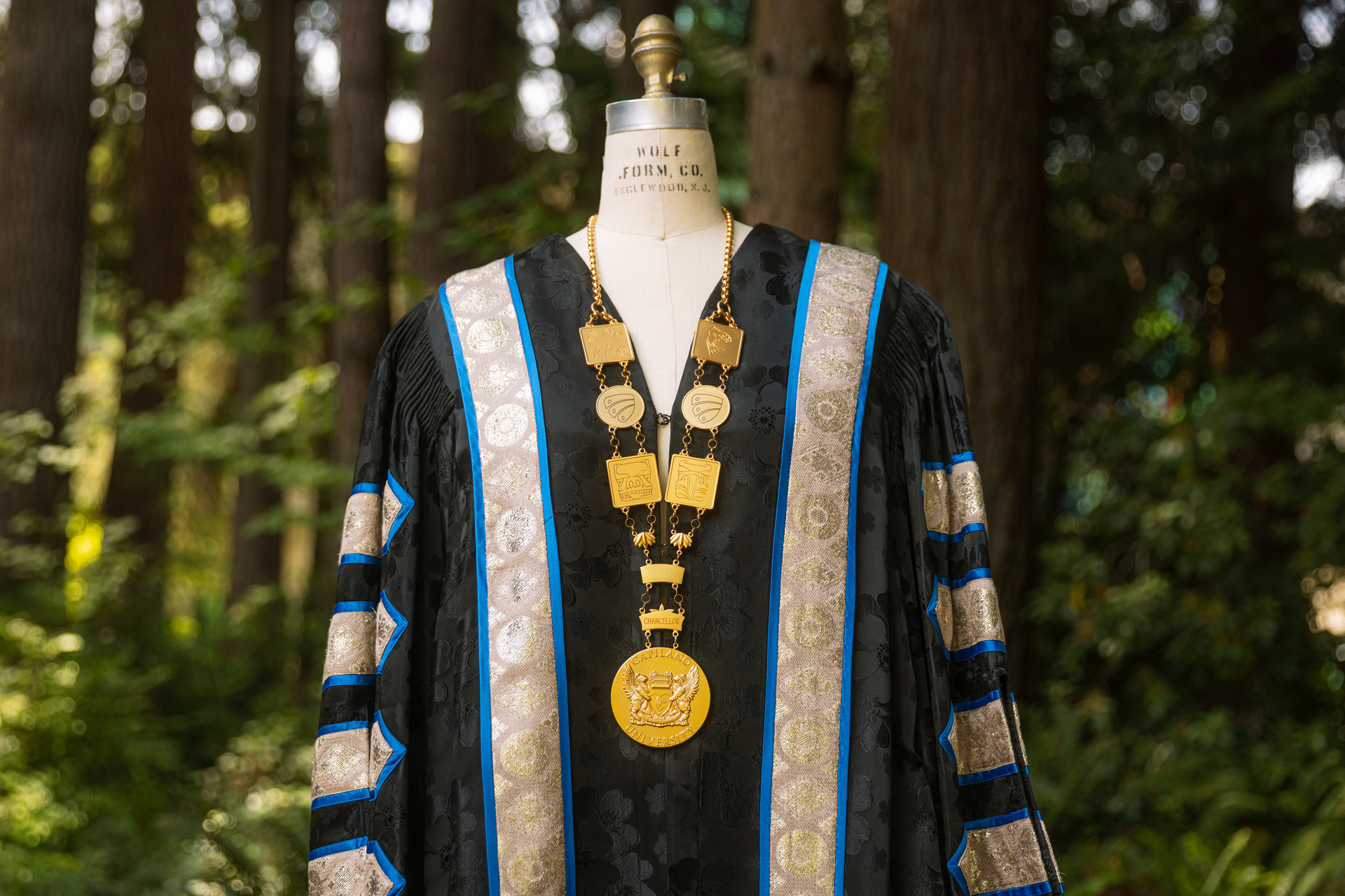 Capilano University Chancellor’s regalia displayed on a mannequin, featuring a black academic gown with blue and silver trim and the official chain of office anchored by a large medallion with the university coat of arms and text reading 