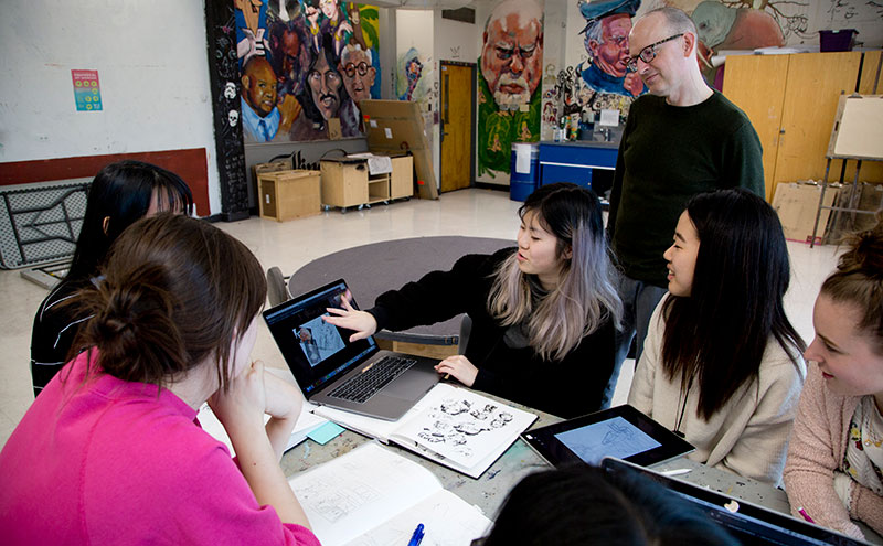 Students sitting at table reviewing design work with instructor watching