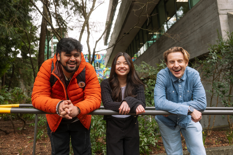 Three students standing side by side, leaning on a railing smiling and facing the camera
